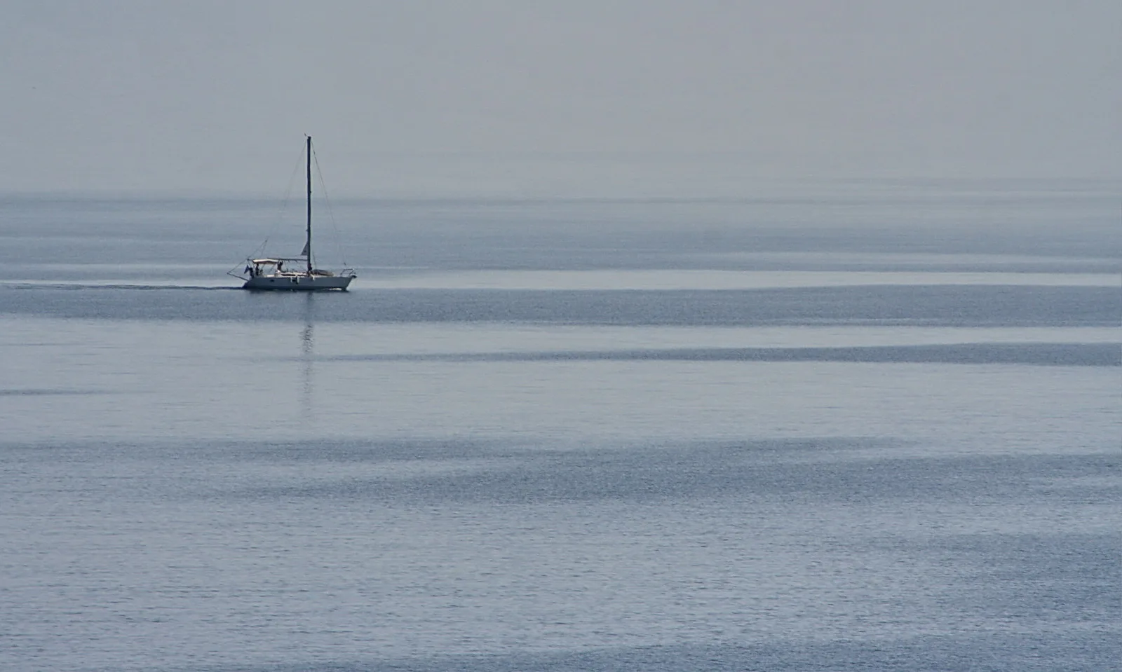 Ein einzelnes weißes Segelboot mit hohem Mast fährt auf einer glatten, grau-blauen Meeresfläche unter bedecktem, dunstigem Himmel.