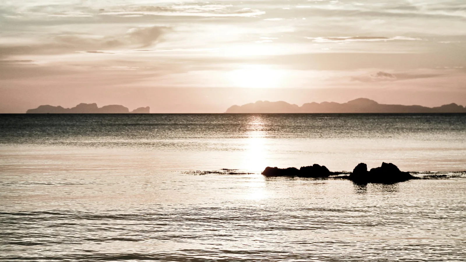 Ruhiges Meer bei Sonnenuntergang, im Vordergrund eine kleine Felsgruppe, am Horizont Silhouetten von Inseln unter einem hellen, dunstigen Himmel.