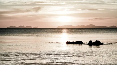 Ruhiges Meer bei Sonnenuntergang, im Vordergrund eine kleine Felsgruppe, am Horizont Silhouetten von Inseln unter einem hellen, dunstigen Himmel.