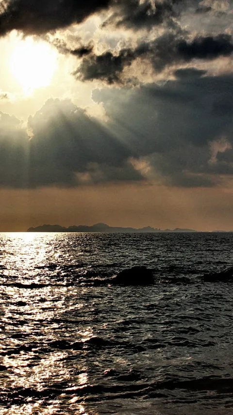 Sonnenstrahlen brechen durch dichte Wolken über einem dunklen Meer, im Vordergrund glitzert die Wasseroberfläche im Gegenlicht, am Horizont zeichnet sich eine flache Inselkette ab.