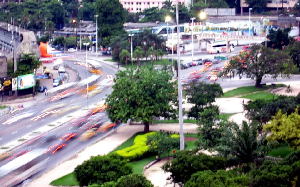 Erhöhte Ansicht einer mehrspurigen Straße in Rio de Janeiro bei Dämmerung, Autos ziehen Lichtstreifen, daneben ein Park mit Bäumen, Palmen und Grünflächen, im Hintergrund Gebäude und eine Brücke.