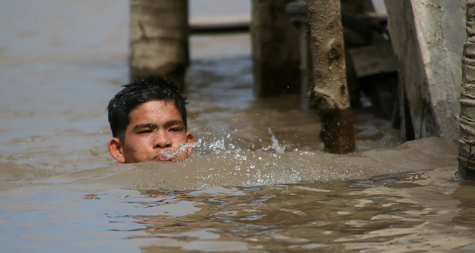 Ein Junge mit nassen dunklen Haaren schwimmt im trüben braunen Wasser, nur Kopf und Schultern ragen heraus. Vor ihm spritzt Wasser, im Hintergrund stehen Holzpfähle im Fluss.