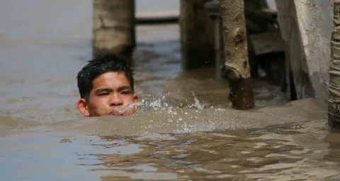 Ein Junge mit nassen dunklen Haaren schwimmt im trüben braunen Wasser, nur Kopf und Schultern ragen heraus. Vor ihm spritzt Wasser, im Hintergrund stehen Holzpfähle im Fluss.