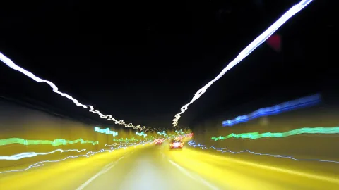 Nächtliche Autobahn aus Fahrerperspektive mit verwischten gelben und grünen Lichtstreifen, dunkler Himmel und zwei rote Rücklichter voraus