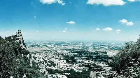 Burg auf einem felsigen Gipfel links im Bild, darunter Dörfer und weite Hügel unter blauem Himmel.