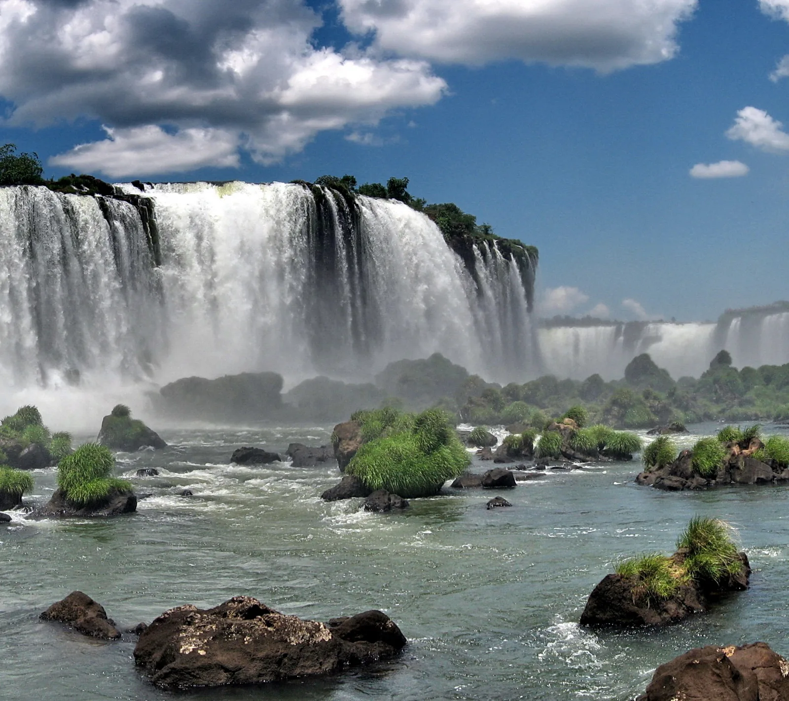 Breiter Wasserfall mit weißer Gischt, davor bewachsene Felseninseln in einem grünlich-grauen Fluss unter blauem Himmel mit Wolken.
