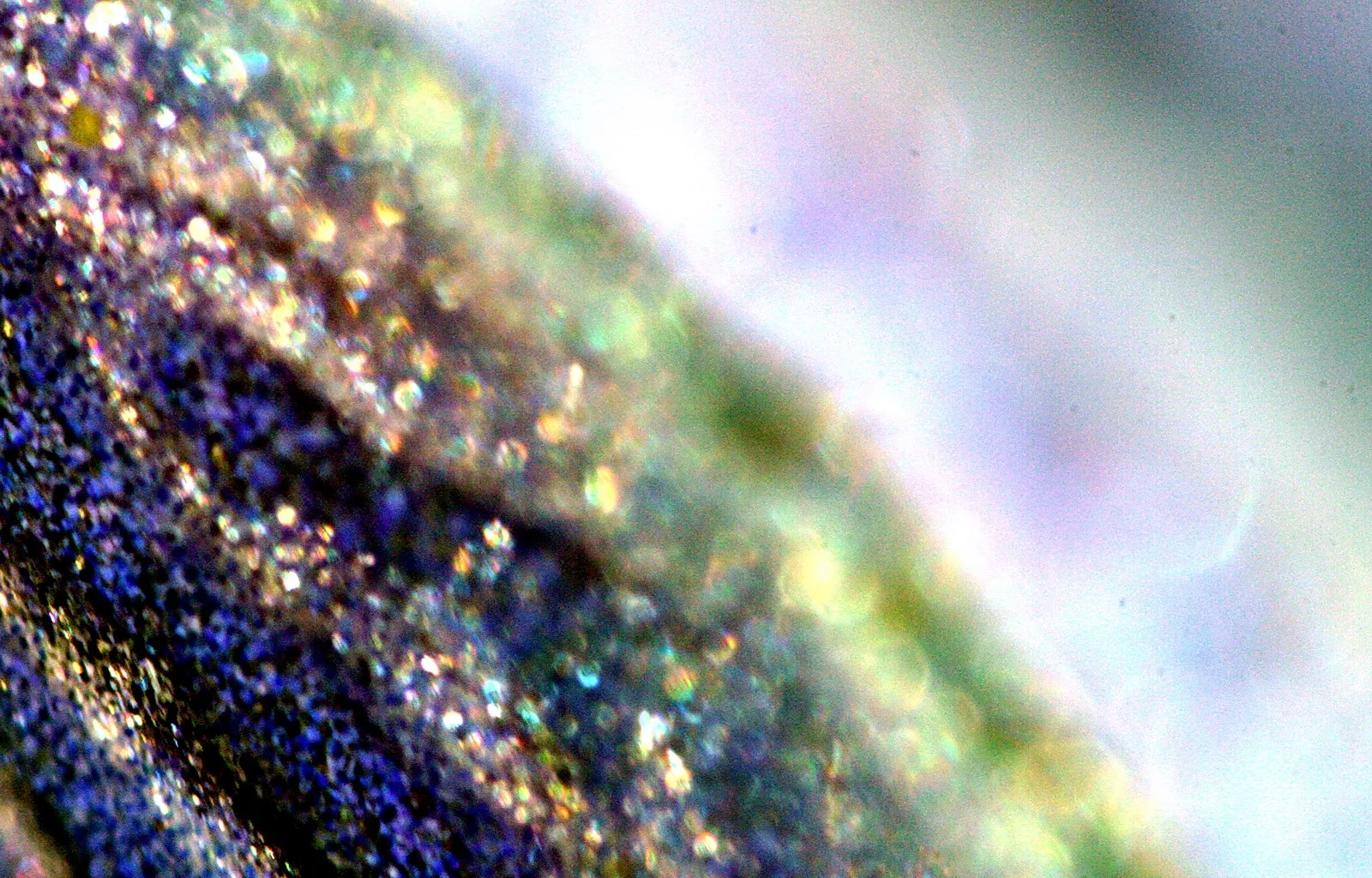 Nahaufnahme einer blau-violetten, körnigen Oberfläche mit schillernden Glitzerpartikeln, die nach rechts in verschwommenes Grün und Lavendel übergeht.