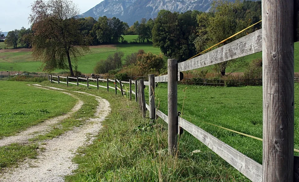 Grauer Holzzaun mit drei Latten läuft entlang eines geschwungenen Kiesweges durch eine grüne Weide, im Hintergrund stehen Bäume und ein Bergrücken.