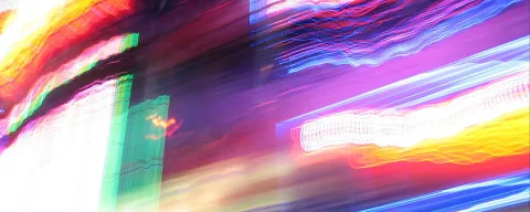 Verwischte Lichtstreifen in Rot, Violett, Blau und Gelb vor dunklem Hintergrund, links ein grüner Leuchtkasten.