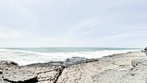 Felsige Küste im Vordergrund, dahinter das Meer mit weißer Brandung unter einem hellen, leicht bewölkten Himmel