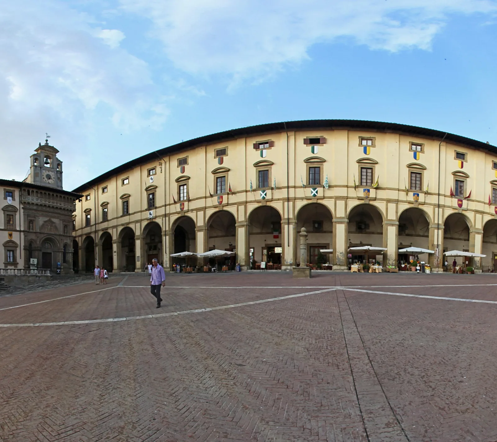 Großer gepflasterter Platz in Arezzo mit einem halbrunden, zweistöckigen Arkadengebäude mit Rundbögen und Sonnenschirmen vor Cafés, links ein Palazzo mit Uhrturm, davor ein Mann im Hemd, der über den Platz geht.