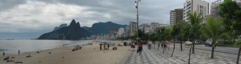 Breiter Sandstrand mit Badegästen am Meer, links eine zerklüftete Bergkette unter bewölktem Himmel, rechts eine Strandpromenade mit Palmen, Passanten, Autos und Hochhäusern.