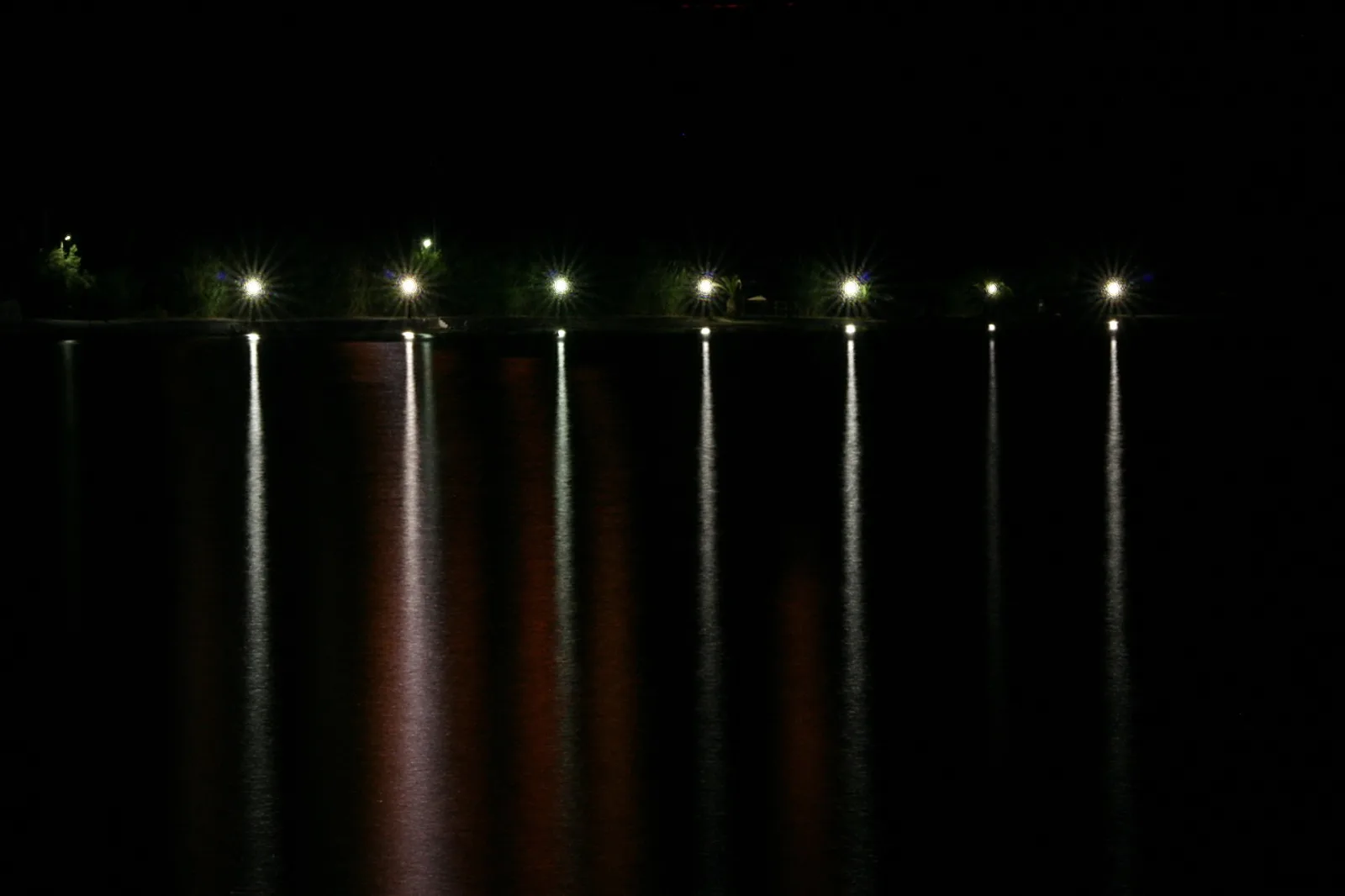 Eine Reihe heller Lampen entlang eines dunklen Ufers, deren Licht sich als senkrechte Streifen im schwarzen Wasser spiegelt.