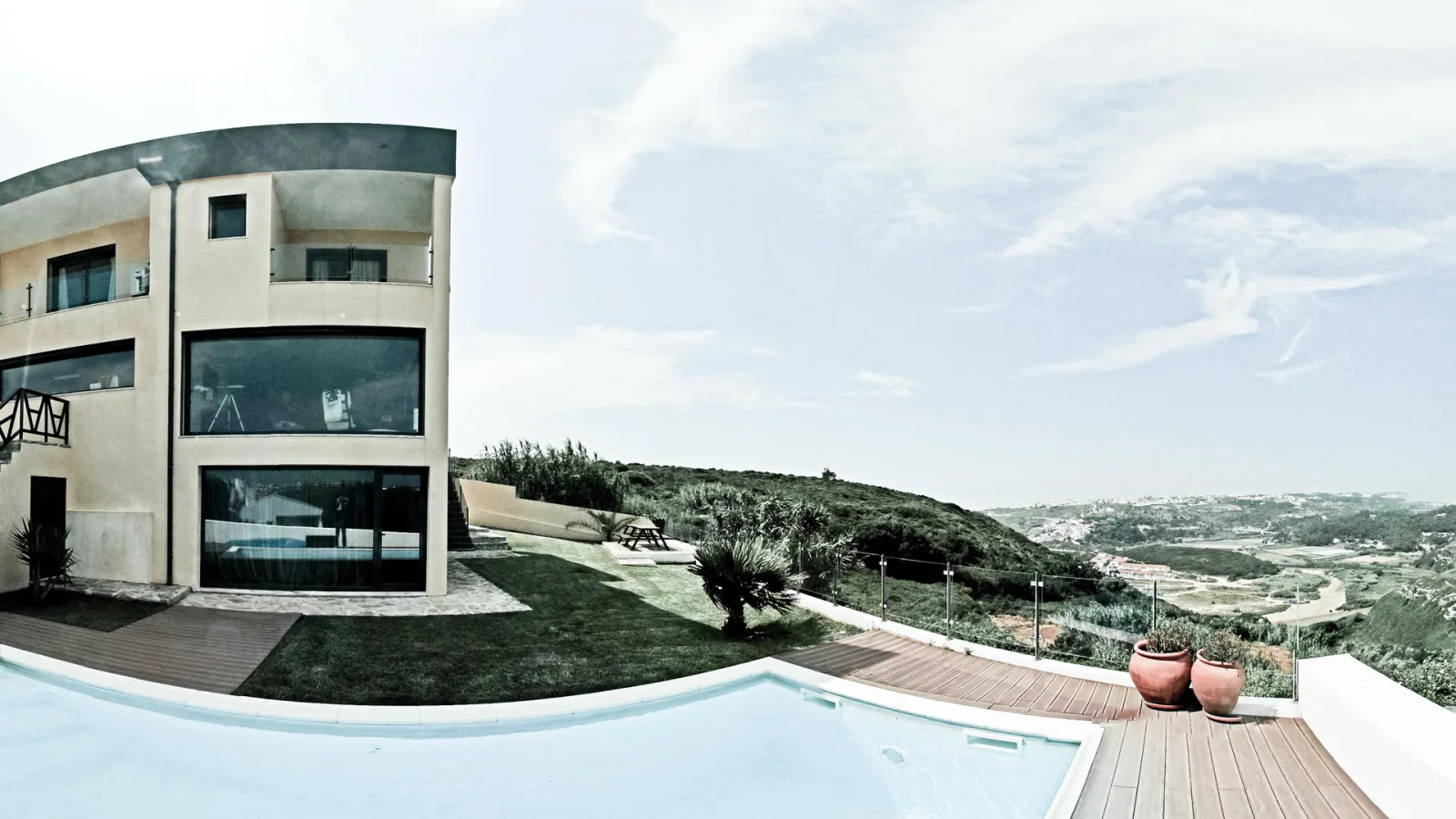 Moderne Villa mit großen Glasfronten, davor ein Infinity-Pool mit Holzdeck, im Hintergrund hügelige Landschaft und Küstenebene unter weitem Himmel.