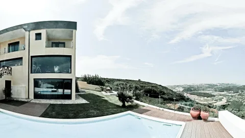 Moderne Villa mit großen Glasfronten, davor ein Infinity-Pool mit Holzdeck, im Hintergrund hügelige Landschaft und Küstenebene unter weitem Himmel.