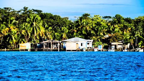 Blaues Wasser im Vordergrund, dahinter eine Reihe einfacher Holzhäuser auf Pfählen am Ufer, umgeben von dichten Palmen und Laubbäumen unter blauem Himmel.