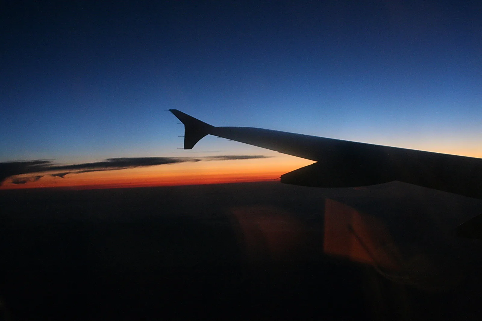 Silhouette eines Flugzeugflügels vor einem Abendhimmel mit orangefarbenem Horizont über einer Wolkendecke.