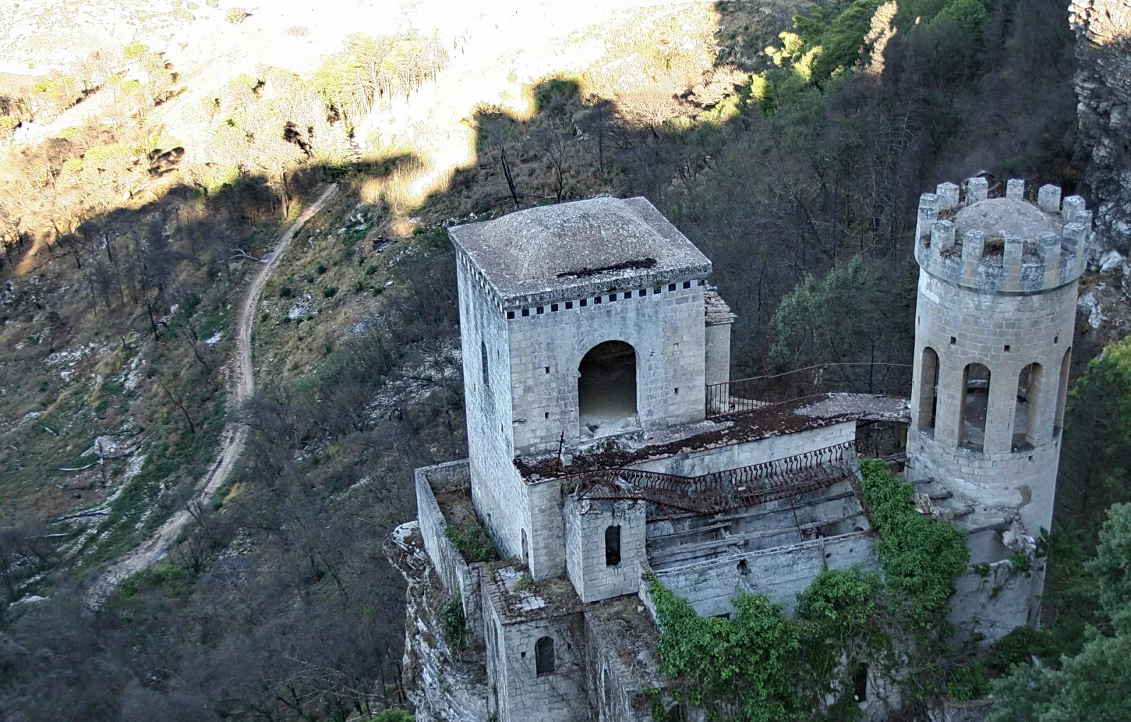 Alte Burg mit Rundturm und Zinnen auf einem Felsvorsprung, davor ein steiler, bewaldeter Abhang mit einem schmalen Pfad.