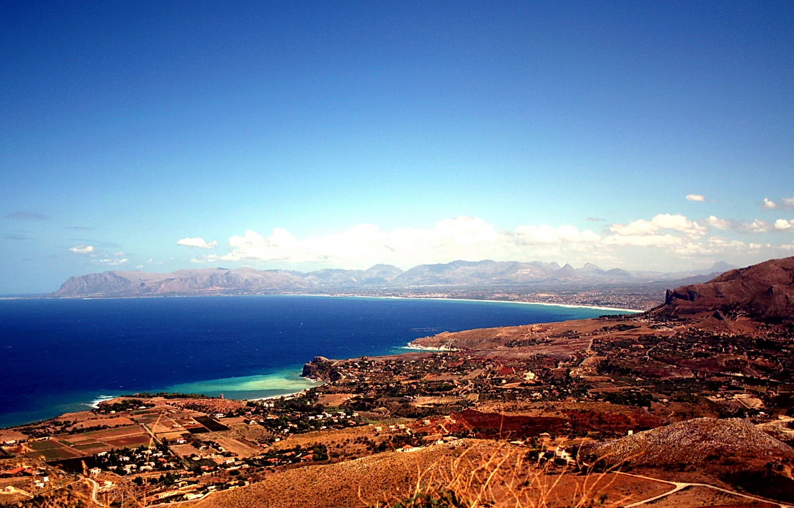 Blick von einer Anhöhe auf eine Bucht mit türkisfarbenem Wasser, einem Küstenort und Bergen unter blauem Himmel.