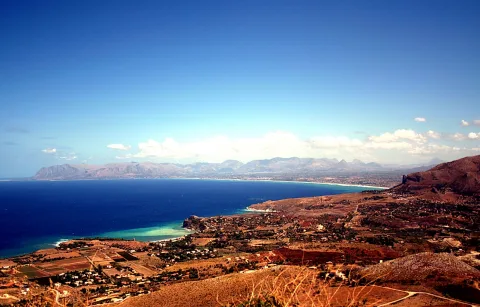 Blick von einer Anhöhe auf eine Bucht mit türkisfarbenem Wasser, einem Küstenort und Bergen unter blauem Himmel.
