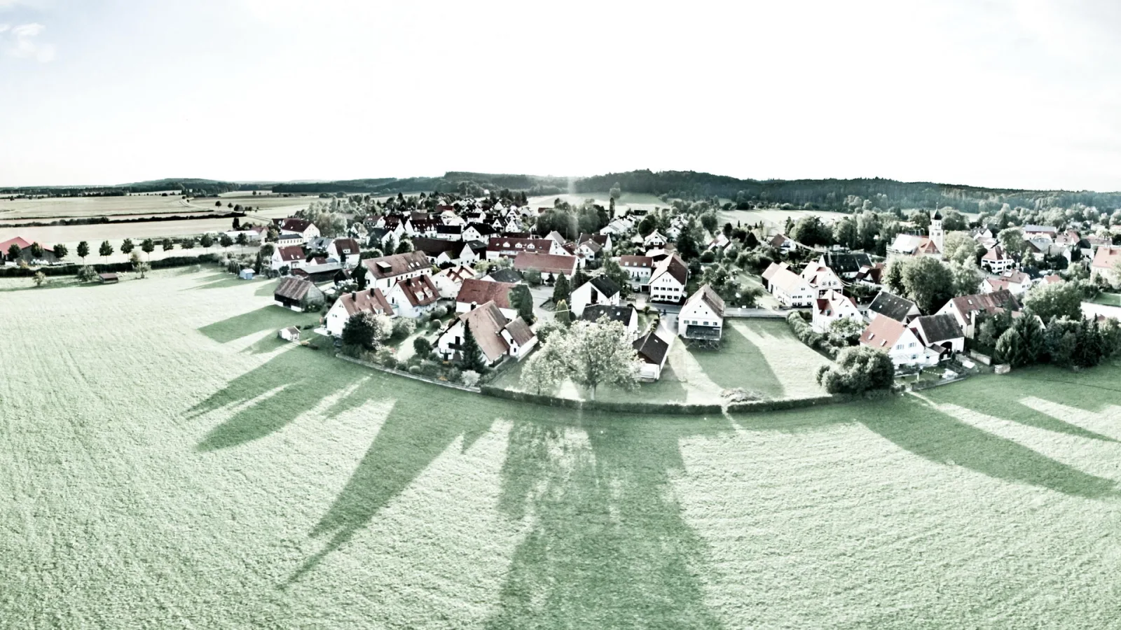 Panoramablick auf ein Dorf mit roten Dächern, Feldern und Wiesen, lange Schatten fallen auf die Wiese im Vordergrund.