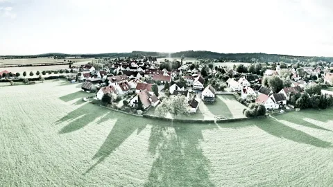 Panoramablick auf ein Dorf mit roten Dächern, Feldern und Wiesen, lange Schatten fallen auf die Wiese im Vordergrund.