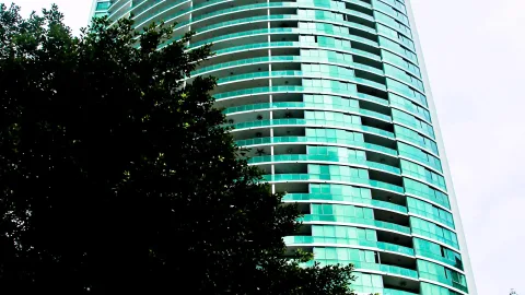 Ein rund geschwungenes Hochhaus mit türkisfarbenen Glasbalkonen, davor links ein dichter dunkler Baum vor hellem Himmel.