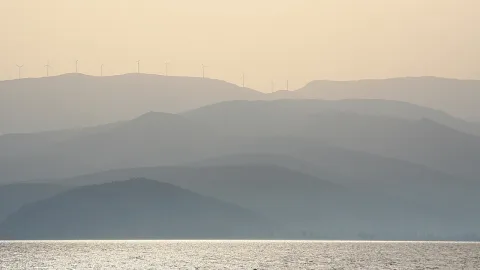 Mehrere Windräder auf einem Bergkamm, davor gestaffelte, dunstige Hügelketten und ein glitzerndes Meer.