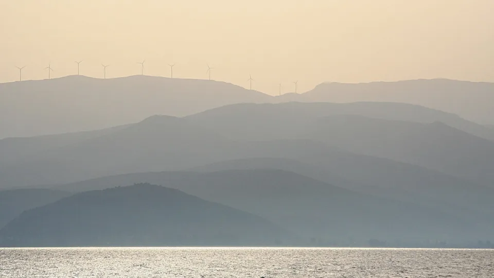 Mehrere Windräder auf einem Bergkamm, davor gestaffelte, dunstige Hügelketten und ein glitzerndes Meer.
