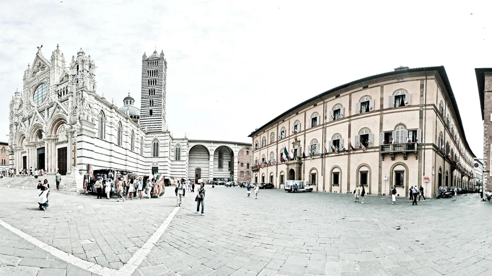 Weiter Platz mit Pflastersteinen in Siena, links die gestreifte Domfassade mit Glockenturm, rechts ein langgestreckter Palast mit Fahnen, davor kleine Menschengruppen.