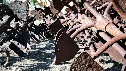Viele verrostete Metallteile landwirtschaftlicher Geräte mit Scheiben und Stangen liegen auf trockenem, steinigem Boden, im Hintergrund grüne Büsche.