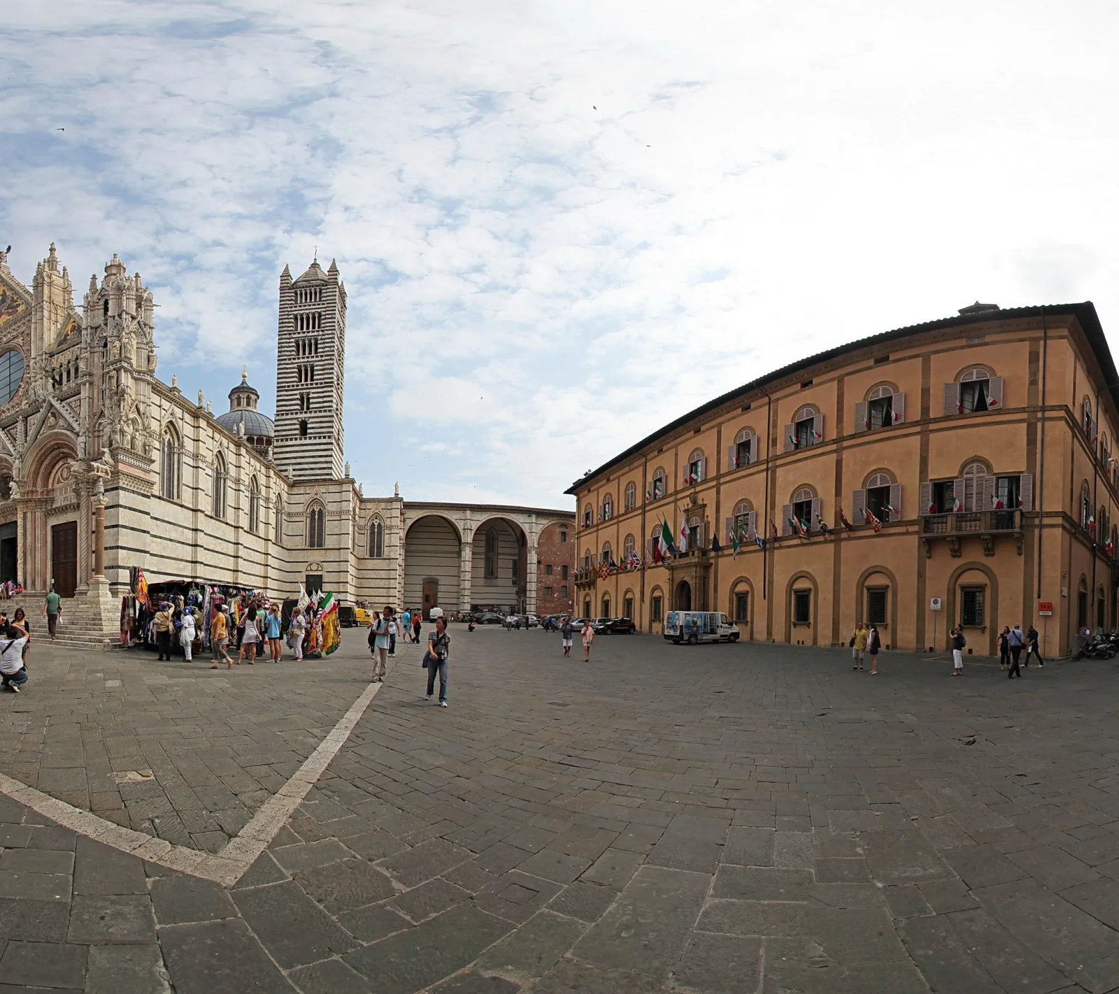 Weiter Platz in Siena mit der hellen, gestreiften Domfassade links, dem Glockenturm und einem ockerfarbenen Palast rechts, davor Touristen und ein Marktstand unter bewölktem Himmel.