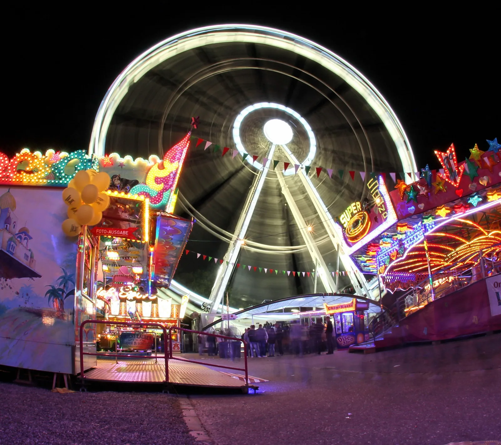 Ein sich drehendes Riesenrad vor dunklem Nachthimmel, davor bunt beleuchtete Fahrgeschäfte und Buden auf einem Jahrmarkt.