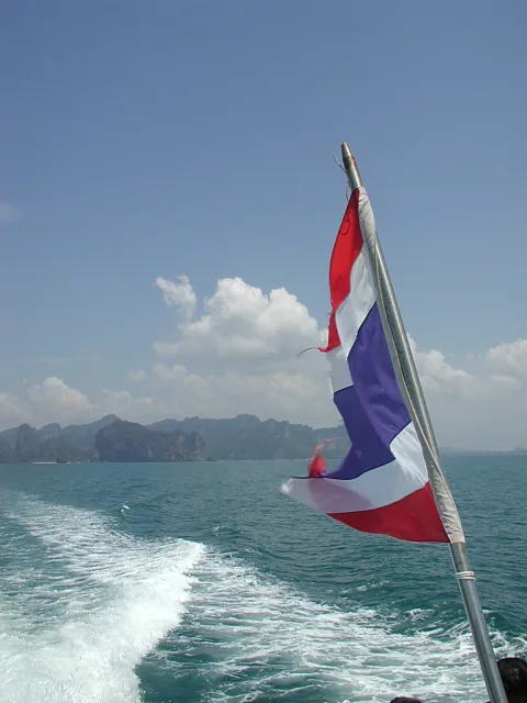 Rot-weiß-blaue thailändische Flagge weht am Heck eines Bootes, dahinter schäumt das Kielwasser und Kalksteinfelsen ragen am Horizont auf.