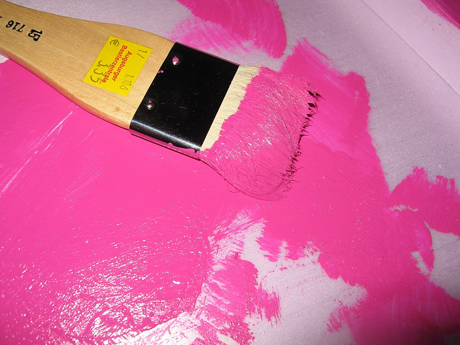 Ein flacher Pinsel mit holzgriff und schwarzer Zwinge liegt auf einer großflächig aufgetragenen, leuchtend pinken Farbschicht, deren Ränder hellgrau durchschimmern.