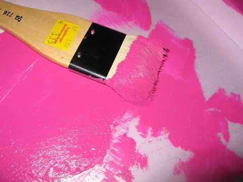 Ein flacher Pinsel mit holzgriff und schwarzer Zwinge liegt auf einer großflächig aufgetragenen, leuchtend pinken Farbschicht, deren Ränder hellgrau durchschimmern.