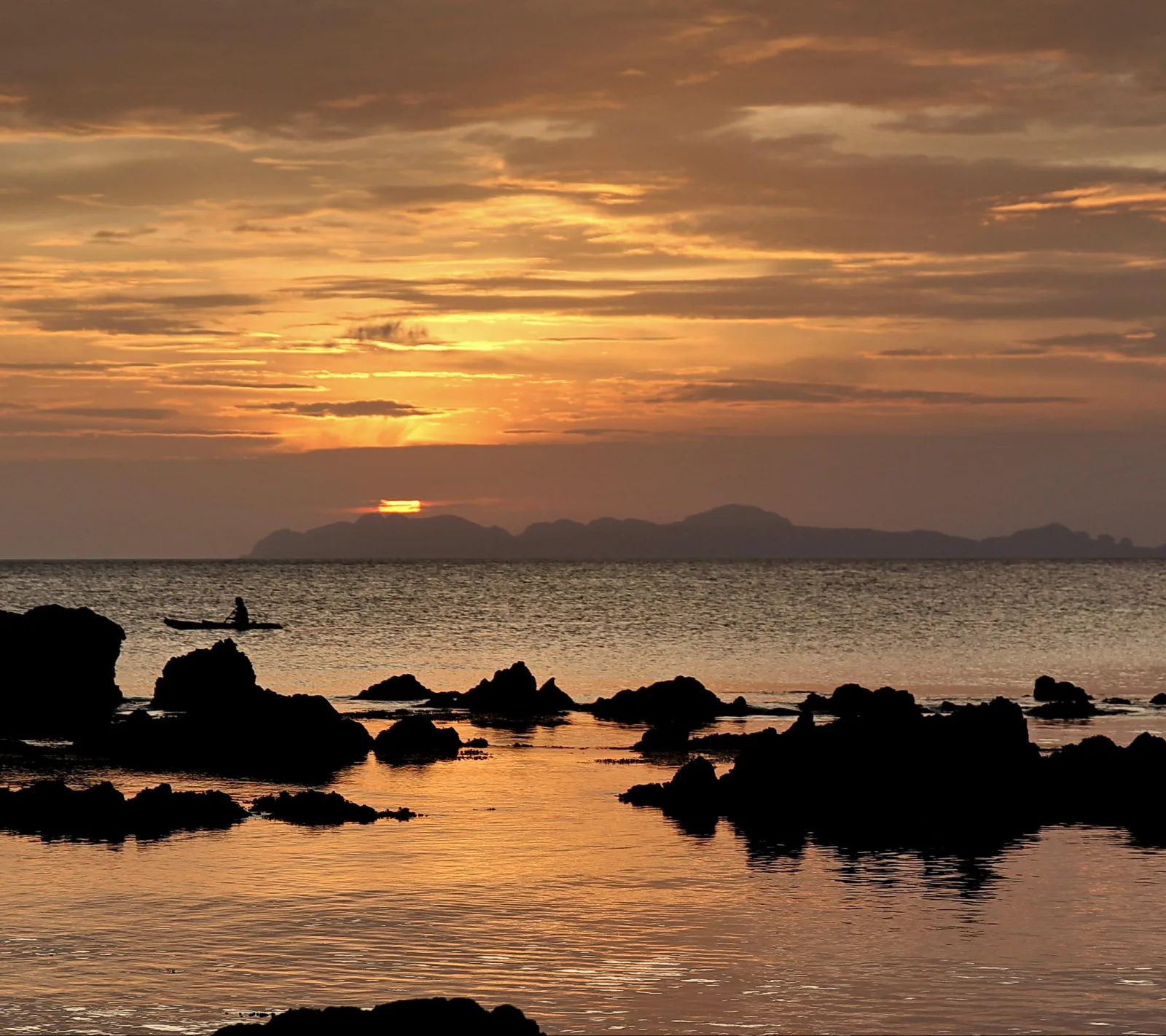 Silhouette eines Kanus mit einer Person auf ruhigem Meer, im Vordergrund dunkle Felsen, dahinter Inseln und eine tief stehende Sonne in orangefarbenem Abendhimmel.