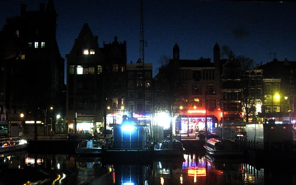 Nächtlicher Blick auf eine Gracht in Amsterdam mit beleuchteten Backsteinhäusern, angelegten Booten und Lichtspiegelungen auf dem Wasser
