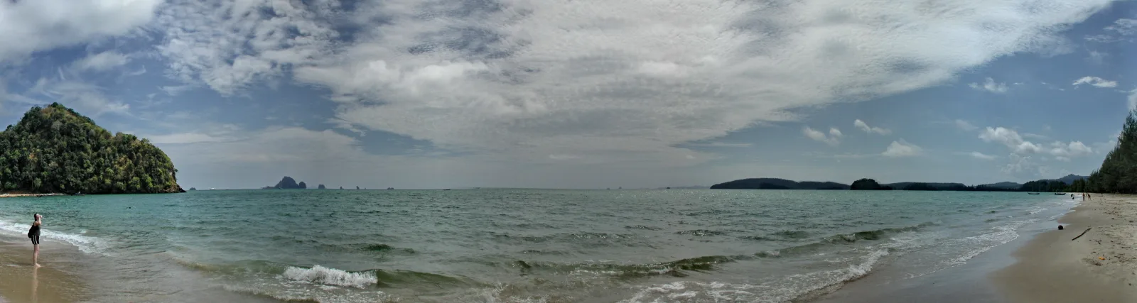 Panoramablick auf einen flachen Sandstrand mit türkisfarbenem Meer, bewaldeter Felsinsel links und fernen Inseln am Horizont; eine Person steht am linken Wasserrand, der Himmel ist von Wolken bedeckt.