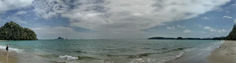Panoramablick auf einen flachen Sandstrand mit türkisfarbenem Meer, bewaldeter Felsinsel links und fernen Inseln am Horizont; eine Person steht am linken Wasserrand, der Himmel ist von Wolken bedeckt.