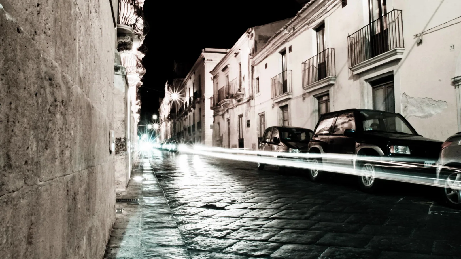 Nächtliche Straße mit Kopfsteinpflaster in einer Altstadt, links eine hohe Steinmauer, rechts barocke Fassaden mit Balkonen und parkende Autos, Lichtspuren vorbeifahrender Fahrzeuge auf dem Asphalt.