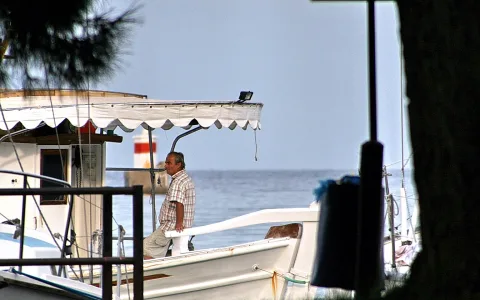 Ein älterer Mann in kariertem Hemd sitzt an Deck eines weißen Fischerboots, dahinter ein Leuchtturm mit rot-weißem Streifen und das ruhige Meer unter klarem Himmel.