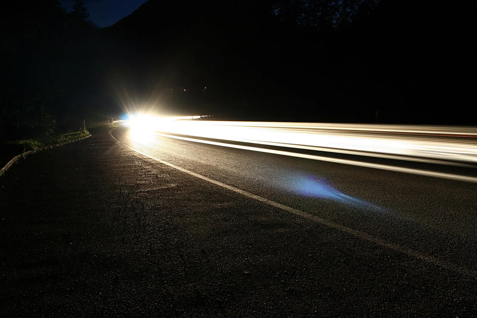 Nächtliche Landstraße, die nach links in eine Kurve führt, mit langen weißen Lichtspuren vorbeifahrender Autos und einem hellen Scheinwerferglanz am Ende der Strecke.