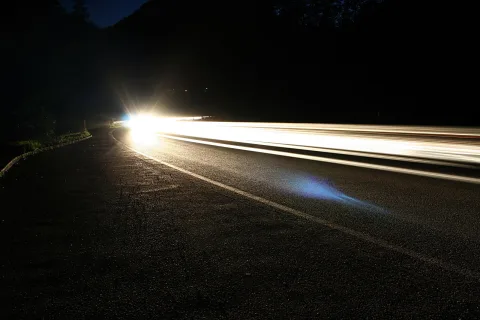 Nächtliche Landstraße, die nach links in eine Kurve führt, mit langen weißen Lichtspuren vorbeifahrender Autos und einem hellen Scheinwerferglanz am Ende der Strecke.