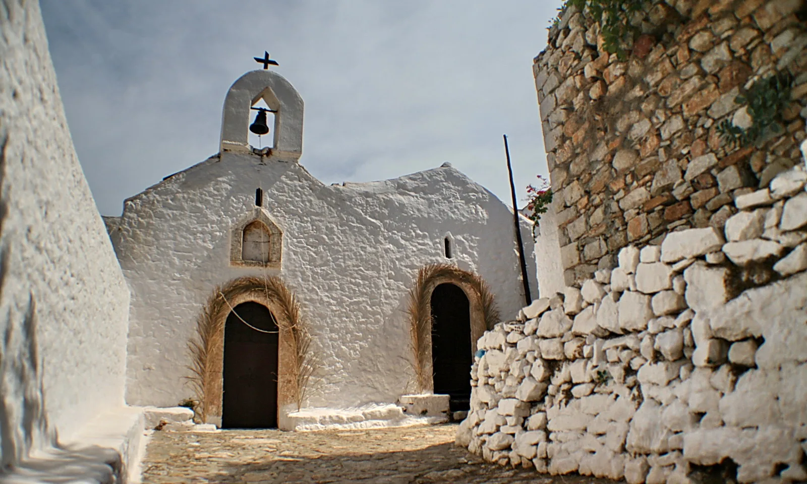 Weiß getünchte Kapelle mit Glockenturm, Glocke und Kreuz auf dem Dach, davor eine Kopfsteingasse, rechts eine hohe Bruchsteinmauer.