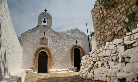 Weiß getünchte Kapelle mit Glockenturm, Glocke und Kreuz auf dem Dach, davor eine Kopfsteingasse, rechts eine hohe Bruchsteinmauer.