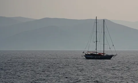 Ein dunkles Segelboot mit zwei Masten fährt auf ruhigem Meer, im Hintergrund ziehen sich mehrere Bergketten im Dunst hintereinander bis zum Horizont.