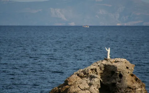 Weiße Jesusstatue mit ausgebreiteten Armen steht auf einem felsigen Vorsprung über dem blauen Meer, im Hintergrund ziehen sich Berge am Horizont entlang, ein kleines Boot fährt vorbei.