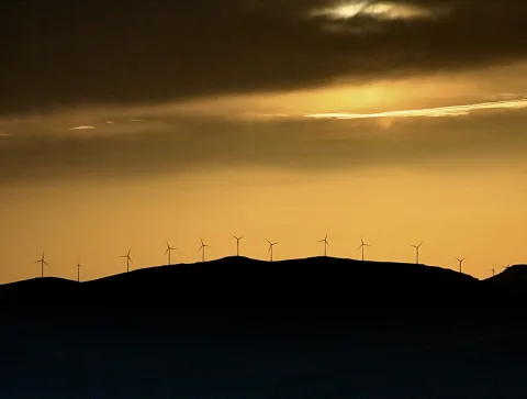 Silhouetten von Windrädern auf einer Hügelkette vor einem goldorangefarbenen Abendhimmel mit dunklen Wolken.