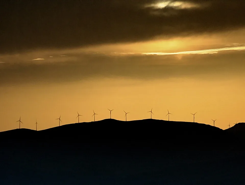 Silhouetten von Windrädern auf einer Hügelkette vor einem goldorangefarbenen Abendhimmel mit dunklen Wolken.